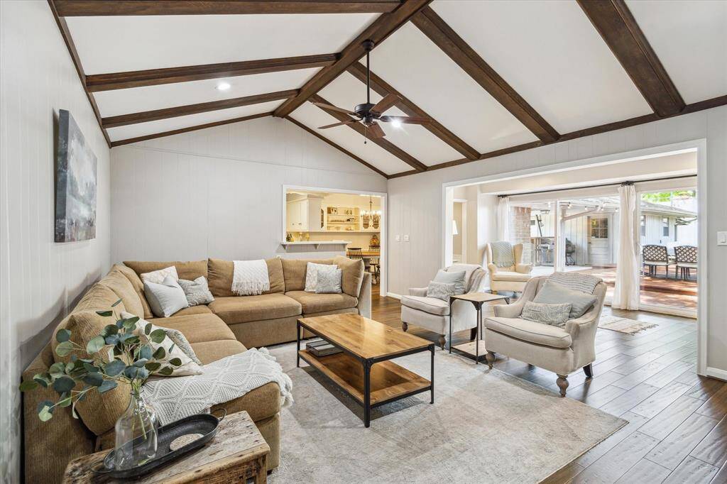 This cozy living room features a vaulted ceiling with exposed wooden beams, a large sectional sofa, and elegant armchairs. The open layout leads to a bright sunroom and outdoor patio, offering ample natural light and a seamless indoor-outdoor flow.
