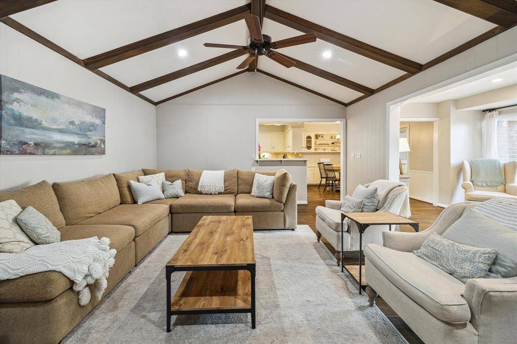This spacious living room features a high ceiling with wooden beams, a cozy sectional sofa, and a warm, neutral color palette. It opens to a bright dining area and kitchen, creating a welcoming and open-concept space.
