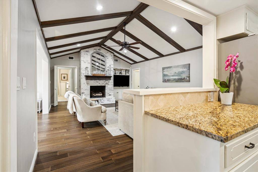 This inviting living space features a vaulted ceiling with exposed wooden beams, a cozy white brick fireplace, and elegant hardwood floors. The open layout connects to the kitchen area with a granite countertop, creating a warm and welcoming ambiance.