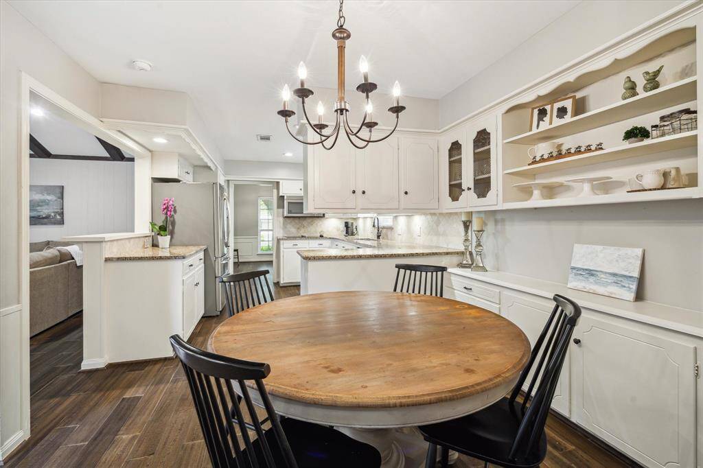 This inviting kitchen features a cozy dining area set against white cabinets with open shelving. The space is bright and airy, with a modern chandelier and stainless steel appliances, seamlessly connecting to the living area.