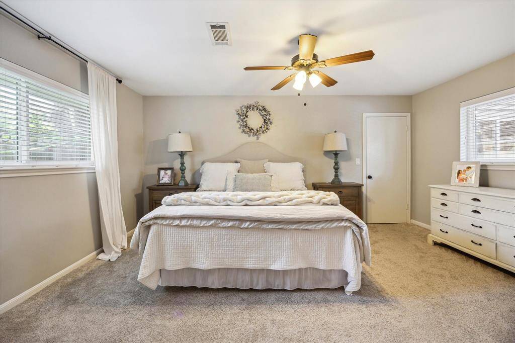 This bedroom is bright and cozy, with neutral tones, plush carpeting, and ample natural light from two windows.