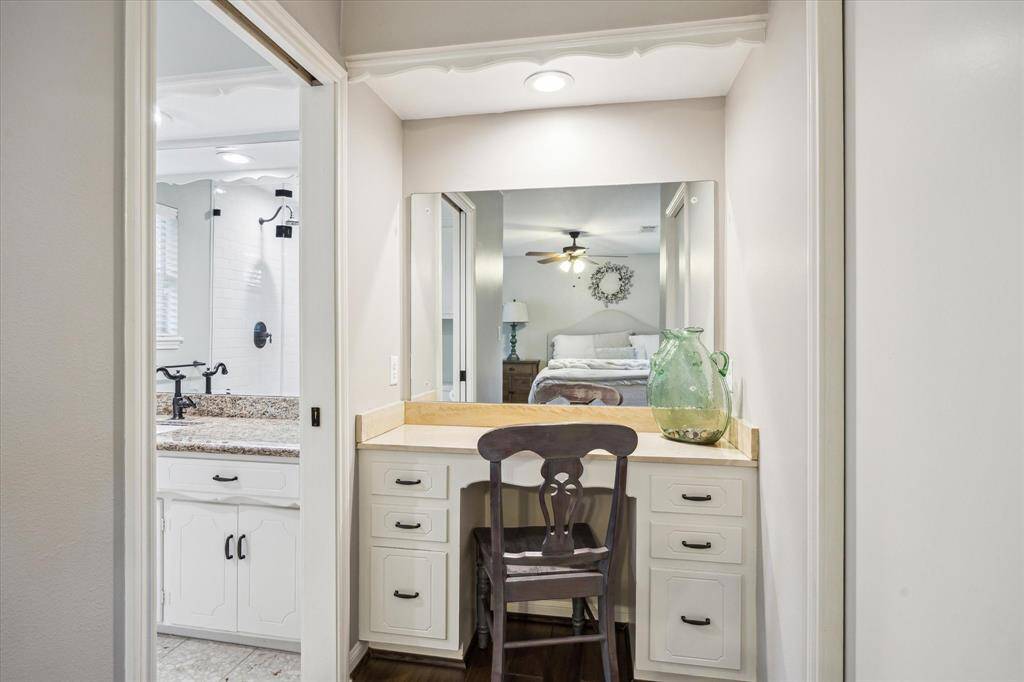 A cozy vanity area with a mirror, chair, and built-in storage, adjacent to a bathroom with white cabinetry and a granite countertop. The space is well-lit and leads into a bedroom, creating a functional and inviting setup.