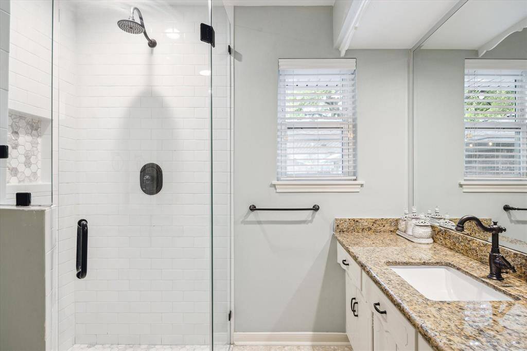 This bathroom features a modern glass-enclosed shower with white subway tiles and a built-in niche. It has dual windows providing natural light, a granite countertop with an undermount sink, and stylish fixtures.