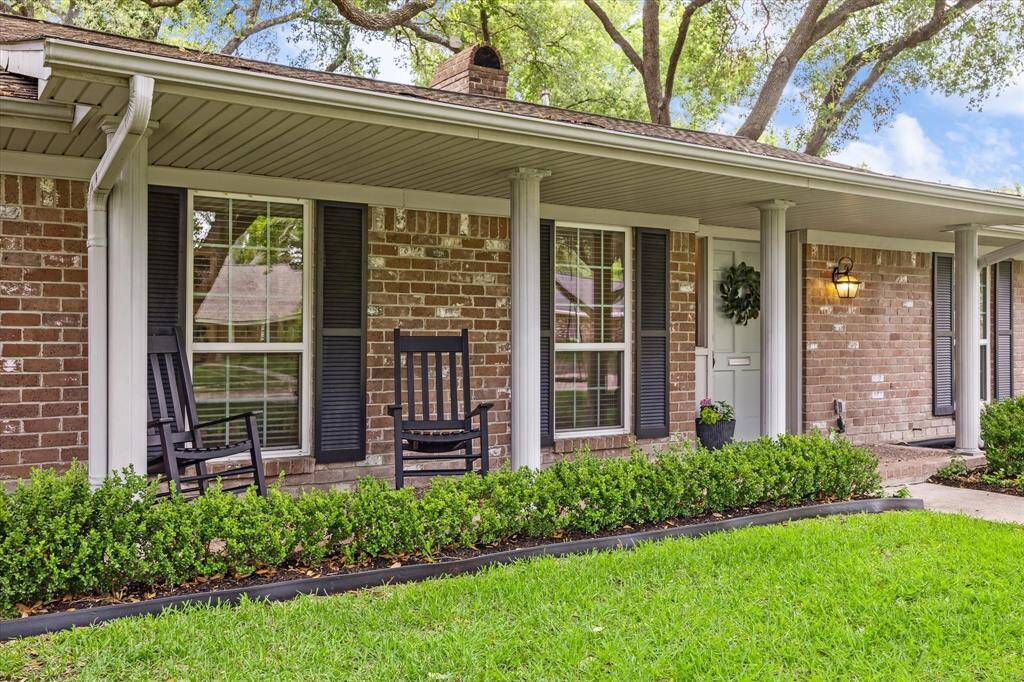 Charming brick ranch-style home with a welcoming front porch, featuring rocking chairs and elegant white columns. The manicured lawn and lush greenery create an inviting atmosphere, perfect for relaxing outdoors.