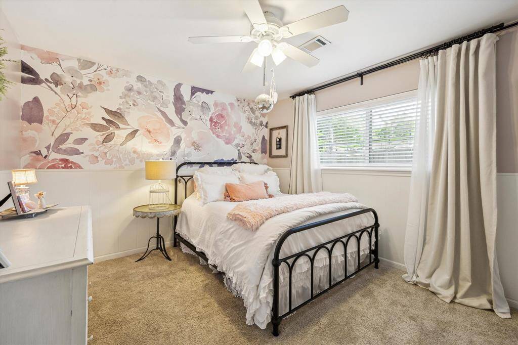This cozy bedroom features a floral accent wall, carpeted floors, and ample natural light from a large window with curtains creating a warm and inviting atmosphere.