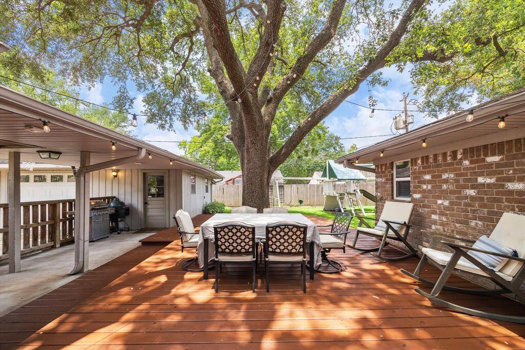This inviting outdoor space features a spacious wooden deck with a dining area under the shade of the large tree. String lights add ambiance, and there's a cozy seating area and grill for entertaining.