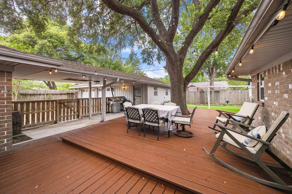 This backyard features a spacious wooden deck with a dining table and chairs, shaded by a large tree. It includes a covered patio area with string lights, perfect for outdoor gatherings.