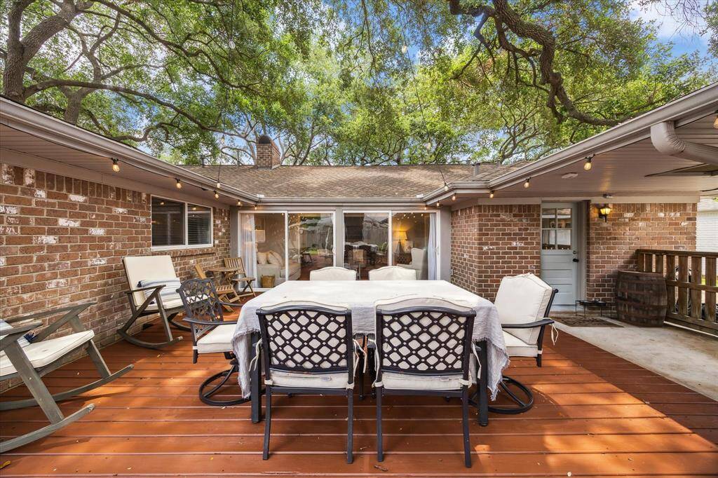 Charming outdoor patio with warm wood decking, surrounded by mature trees. It features comfortable seating, a dining area, and string lights, perfect for relaxation and entertaining. The brick exterior and large sliding doors offer a cozy and inviting atmosphere.