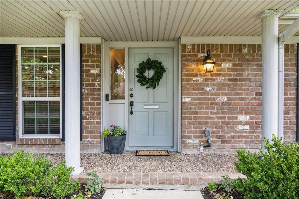 This charming entrance features a welcoming light blue door flanked by classic brickwork and white columns. A cozy porch light adds warmth, and a potted plant provides a touch of greenery.