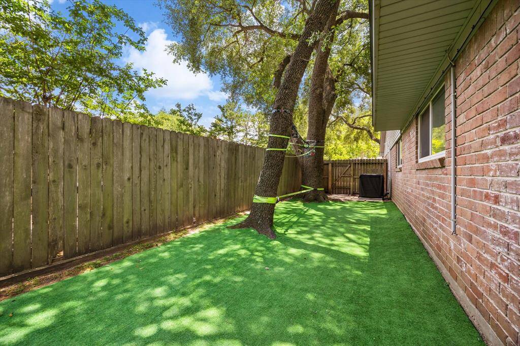 This photo showcases a narrow side yard with artificial grass, a wooden privacy fence, and mature trees providing shade.