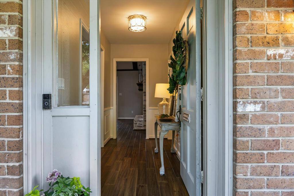 Welcoming home entrance with a brick exterior and a partially open door revealing a bright hallway. The hallway features wood flooring, a decorative table with a lamp, and greenery, creating a cozy and inviting atmosphere.