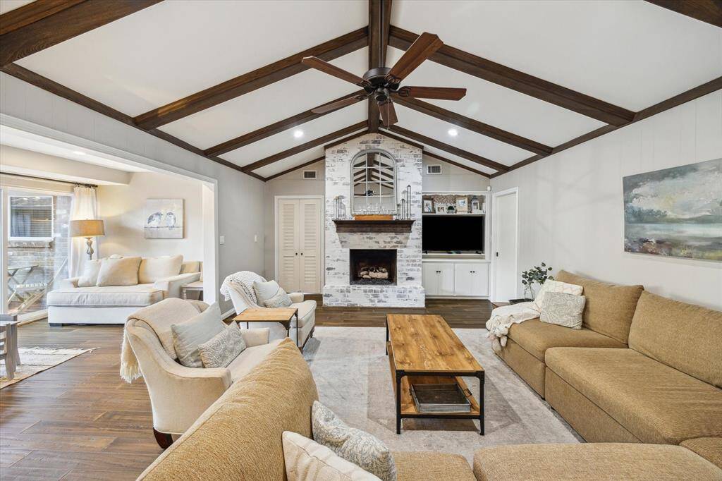This inviting living room features a vaulted ceiling with exposed wooden beams, a cozy brick fireplace, and ample seating with a large sectional sofa. A ceiling fan and natural light from the adjacent sunroom create a warm, welcoming atmosphere.