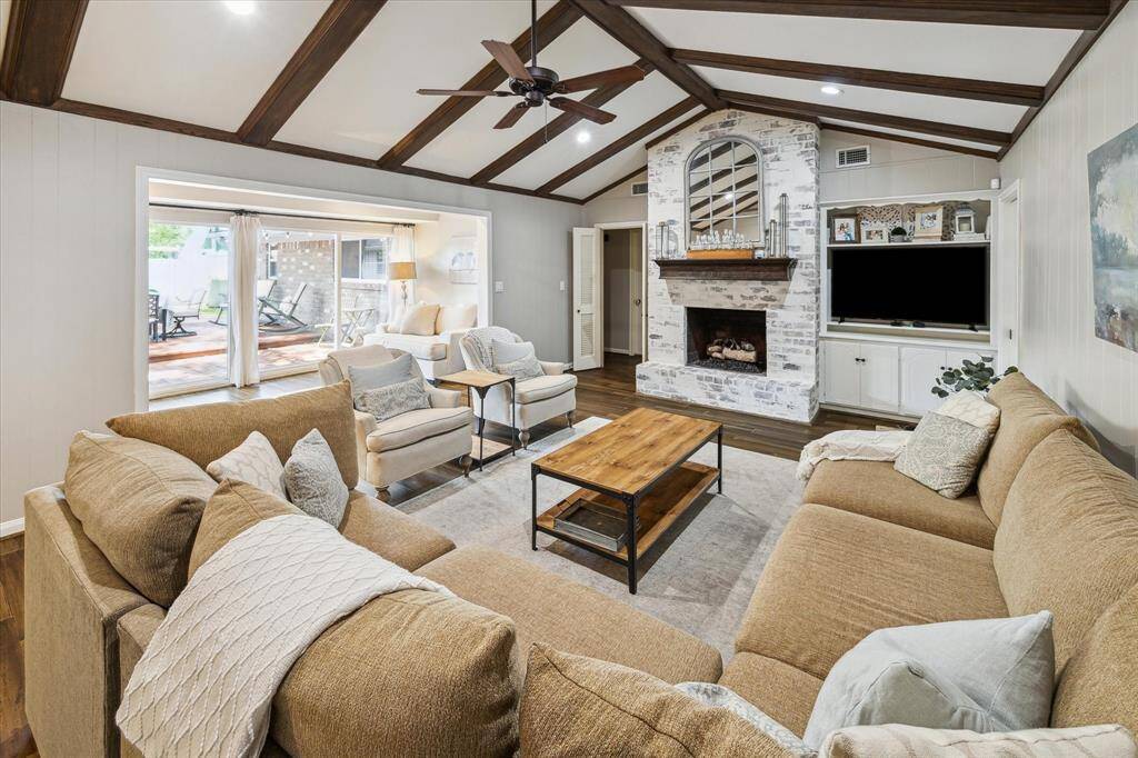 This inviting living room features a vaulted ceiling with exposed beams, a charming brick fireplace, and a spacious layout. It opens to a bright outdoor area, perfect for entertaining. The cozy seating and neutral tones create a warm, welcoming atmosphere.
