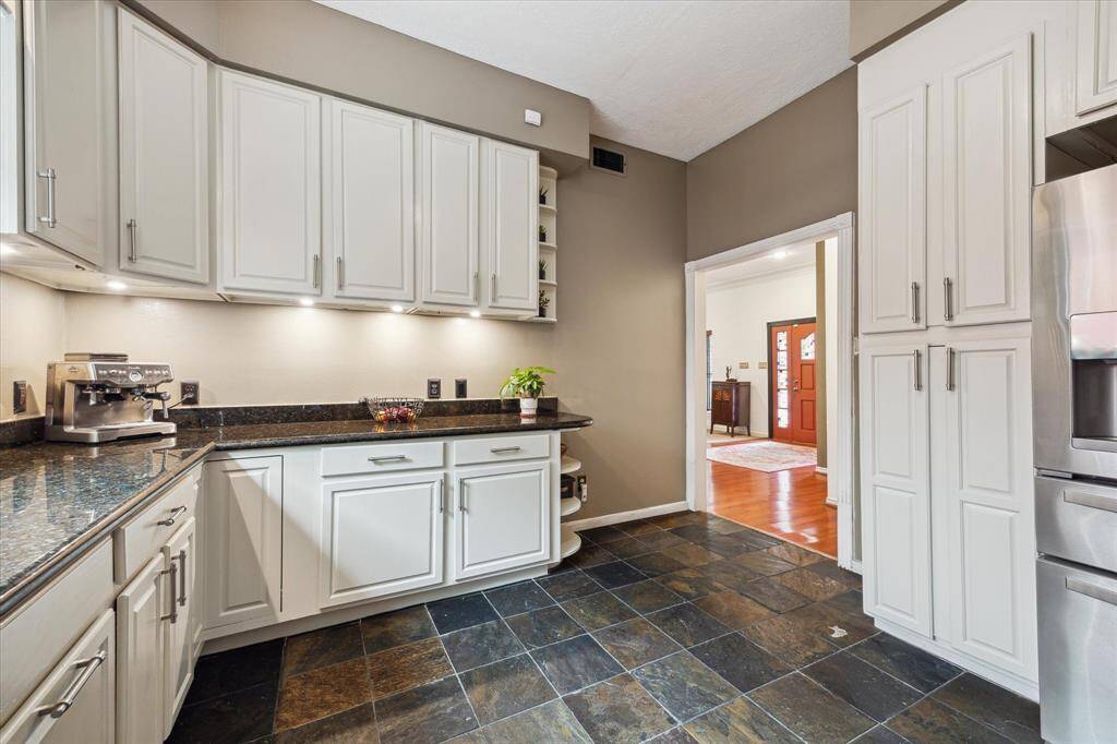 Rear House: Another view of the kitchen features sleek white cabinetry with under-cabinet lighting, a dark granite countertop, and modern stainless steel appliances. It has a slate tile floor and opens into a warmly lit adjoining room with hardwood flooring.
