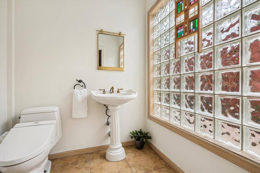 Rear House: Half bathroom features a classic pedestal sink with gold fixtures, a modern toilet, and a glass block window providing natural light and privacy. The neutral tile floor complements the clean, bright aesthetic.