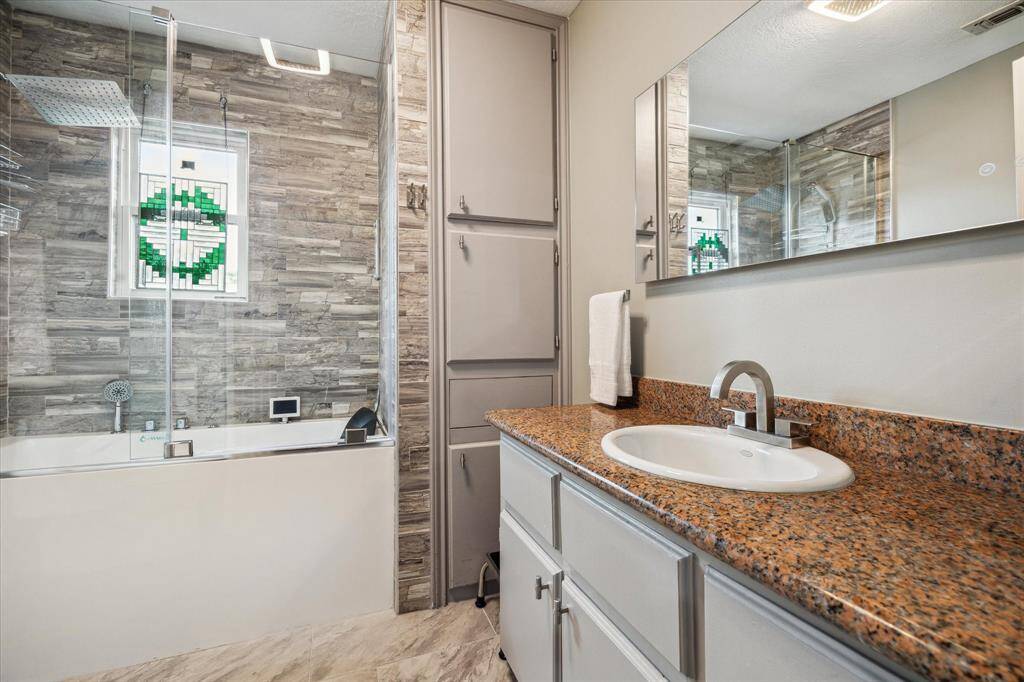 Rear House: Full secondary bathroom features a modern design with a glass-enclosed tub and shower, stone-textured wall tiles, and a granite countertop with an undermount sink. A large mirror and ample storage enhance functionality, while a small stained glass window adds a touch of elegance.