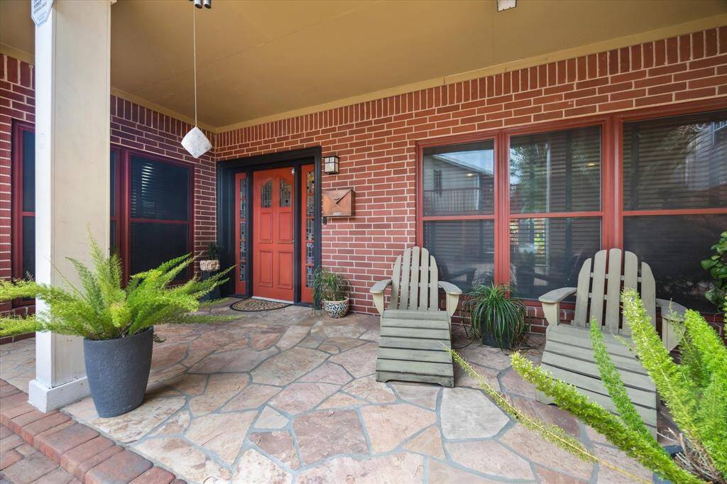Rear House: Front porch of with a warm, inviting red door and brick exterior. Features stone flooring, cozy Adirondack chairs, and lush potted plants, perfect for relaxing.