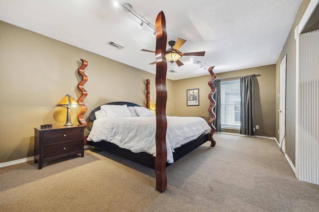 Rear House: Another view of this spacious primary bedroom features neutral walls, and cozy carpeted floor. It has ample natural light from a large window, complemented by modern track lighting and a ceiling fan for comfort.