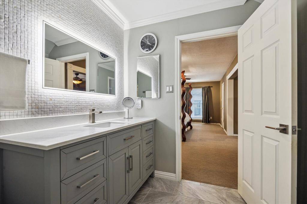Rear House: Another view of the primary bathroom showcasing a sleek double vanity and modern fixtures, complemented by a backlit mirror and mosaic tile wall. The room leads into a well-lit bedroom, offering a seamless transition between spaces.