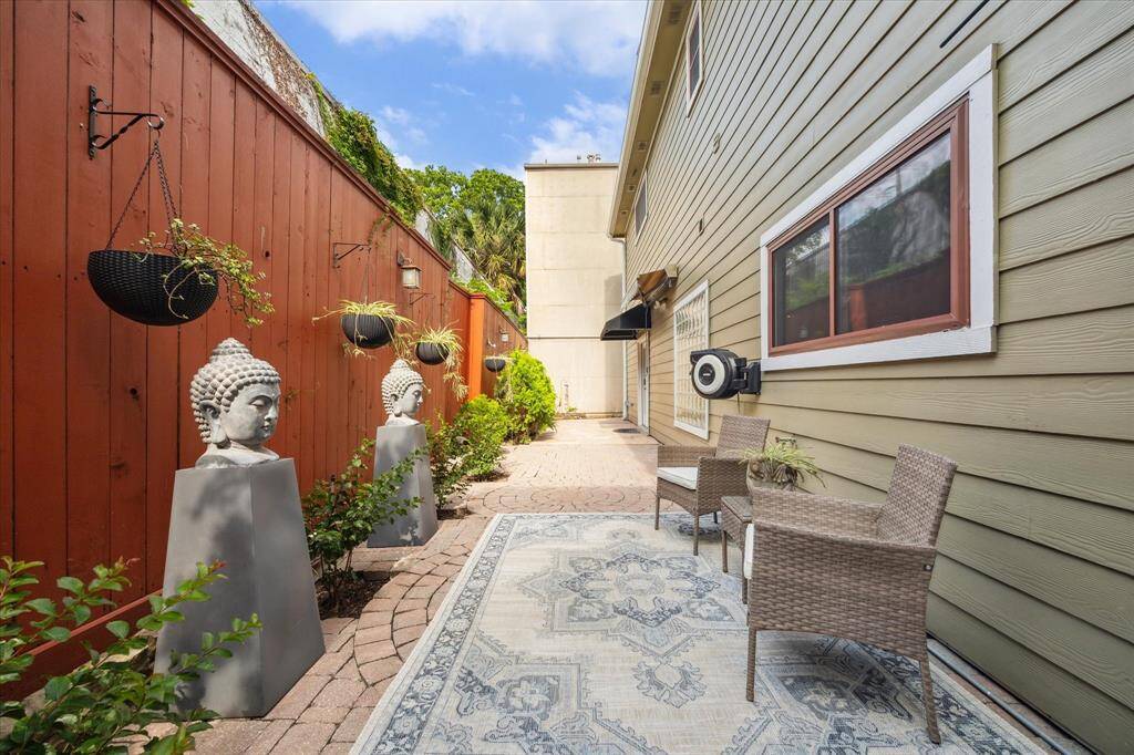 Rear House: Charming outdoor patio space with a brick pathway, decorative statues, hanging plants, and wicker seating. The area is bordered by a warm wood fence and the side of the house, offering a cozy and serene atmosphere.
