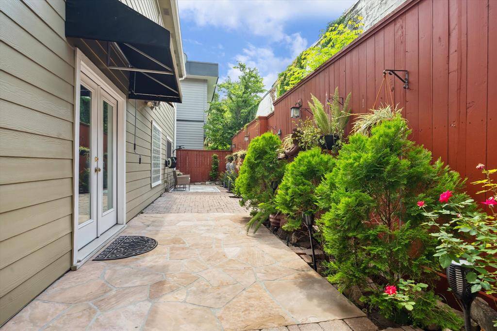 Rear House: A third view of this charming outdoor patio area with stone paving, lush greenery, and a wooden privacy fence. French doors with a black awning lead out from the house, creating a cozy, inviting space perfect for relaxation or entertaining.