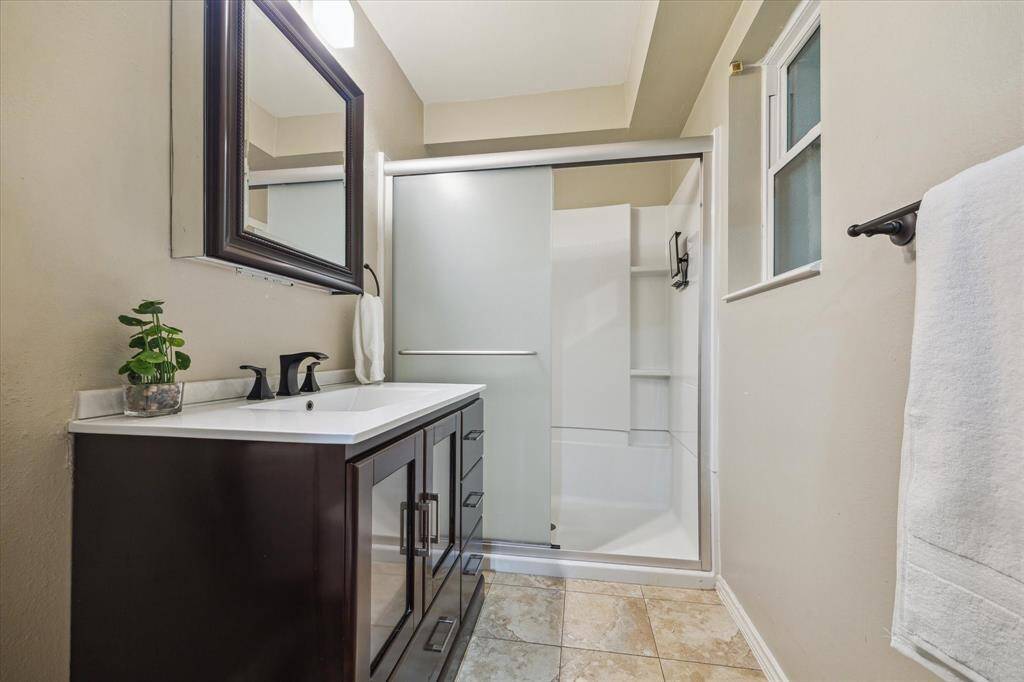Front House: Full downstairs bathroom features a modern vanity with a dark finish and a large mirror. It includes a spacious walk-in shower with frosted glass doors, a small window for natural light, and neutral tile flooring. A towel rack is conveniently placed near the shower.