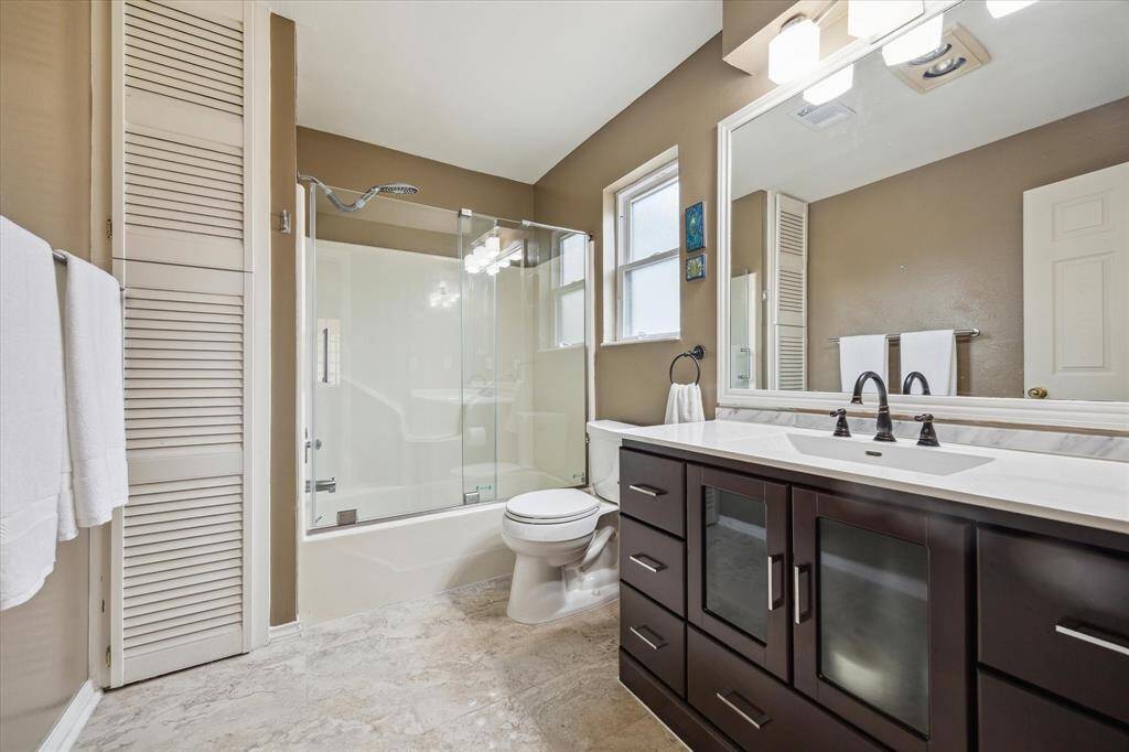 Front House: Full bathroom upstairs has dual sinks, a glass-enclosed bathtub with a shower, and a neutral color palette. It includes a large mirror, ample storage, and natural light from the window, creating a spacious and inviting atmosphere.