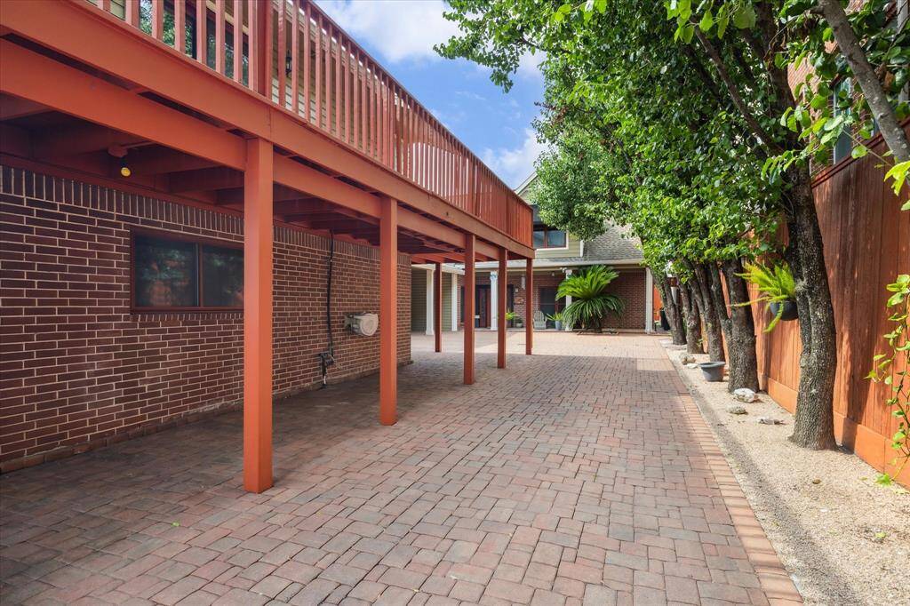 Inlaid brick-paved driveway with a wooden deck above, bordered by trees and a brick wall, offering a private and versatile outdoor space.