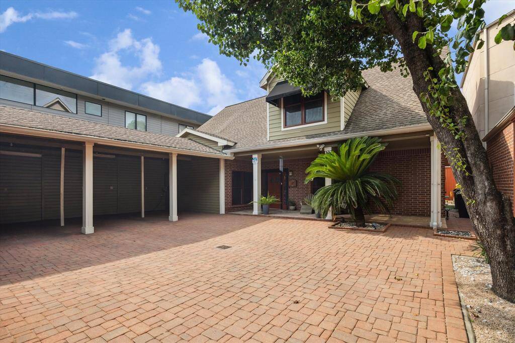 Inlaid brick courtyard features a covered carport and mature landscaping, including a large tree and lush plants, providing a welcoming and private outdoor space.