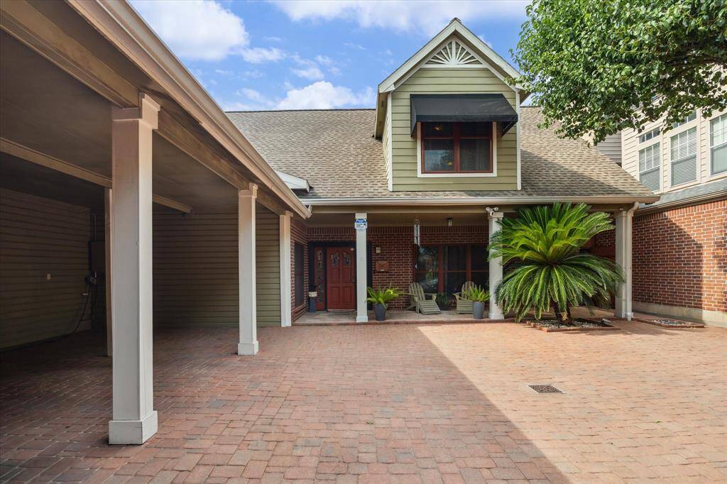 View of rear house with a covered porch and a spacious paved courtyard. Lush greenery and a mature tree provide a welcoming touch. Ideal for relaxing outdoors or entertaining guests.