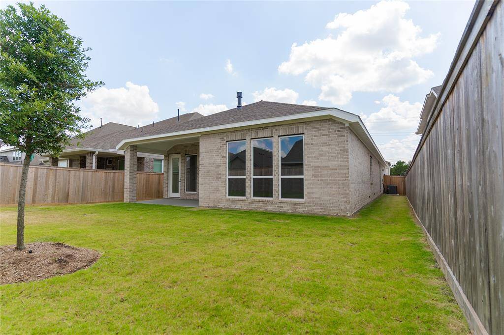 This photo shows a backyard with a well-maintained lawn and a small tree, surrounded by a wooden fence. The house features a brick exterior with large windows and a covered patio, providing a private outdoor space.