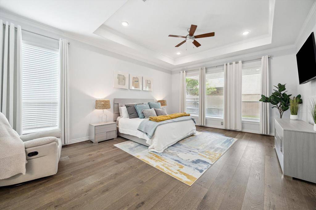 Generous proportions and oversized windows make this primary retreat feel bright and serene. Tray ceilings with recessed lighting add architectural depth, while wide-plank wood flooring brings warmth to the space. A wall of windows filters in soft natural light and offers a view of the backyard, creating a calm and airy atmosphere perfect for unwinding.