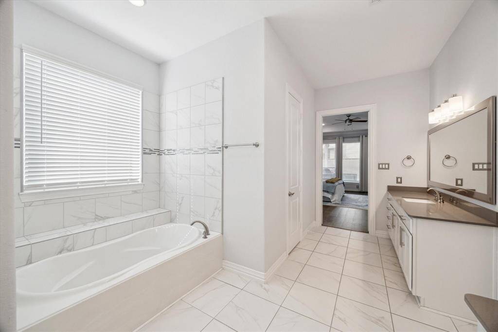 A deep soaking tub wrapped in marble-look tile invites relaxation, framed by a ledge for candles, plants, or bath essentials. Positioned across from the dual vanities, this space balances luxury with everyday comfort. The seamless connection to the primary suite enhances both flow and functionality.