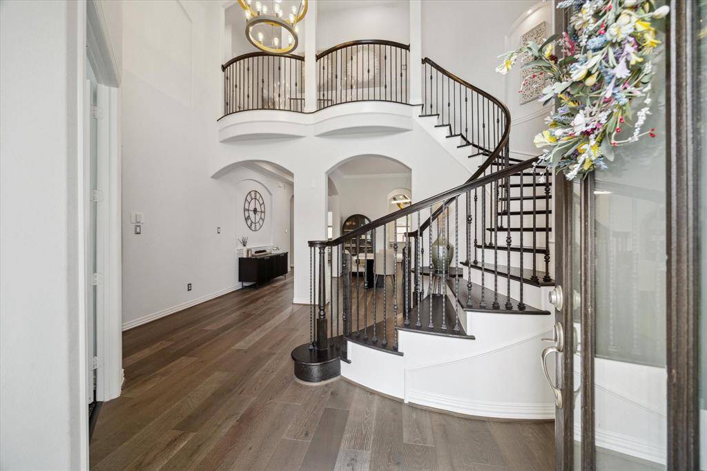 Step through the front door into a grand two-story entryway where architectural elegance takes center stage. A curved staircase with wrought iron spindles adds a striking focal point, complemented by warm wood floors and soaring ceilings. Arched openings lead to the formal dining area and beyond, while a statement chandelier anchors the space with upscale charm and sophistication.