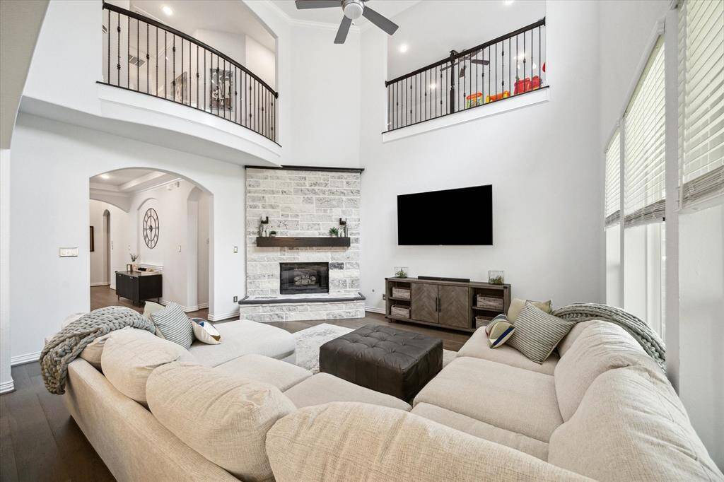 Anchored by a striking floor-to-ceiling stone fireplace, this living area blends grandeur and comfort with its open two-story design and elegant iron railing above. Natural light pours in through oversized windows, highlighting the rich wood floors and architectural archways that connect seamlessly to the surrounding rooms. Ideal for both everyday lounging and entertaining on a larger scale.