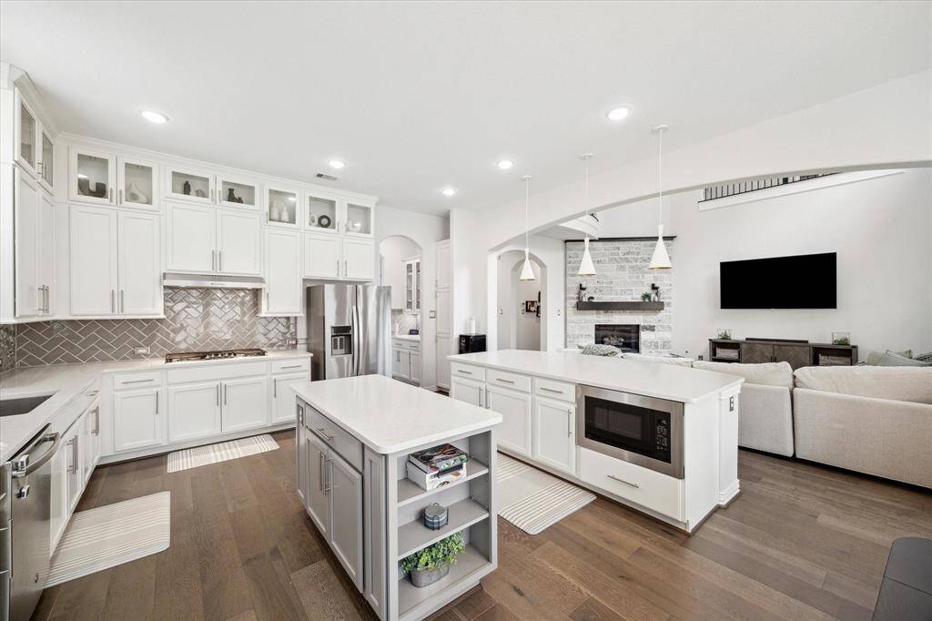 Sleek quartz countertops, soft-close cabinetry, and a stylish herringbone backsplash set a polished tone in this chef-inspired kitchen. Dual islands offer abundant prep space and storage, while the built-in microwave and open shelving add both function and flair. Framed by arched openings, the space connects directly to the living room—perfect for hosting or keeping connected during daily routines.