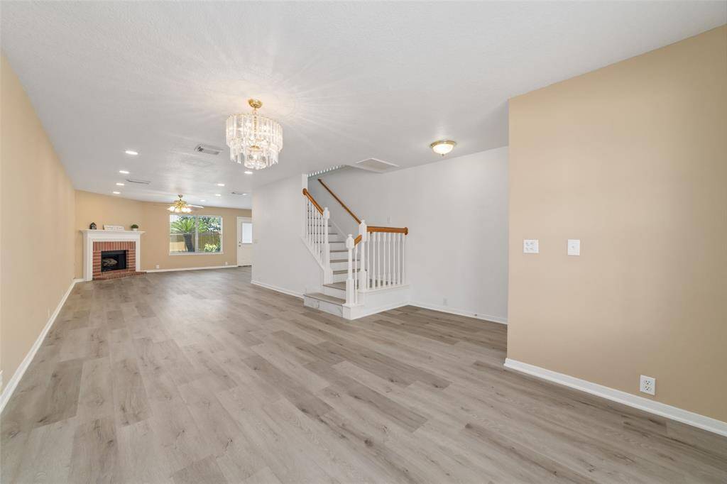 This spacious living area features light laminate flooring, a central chandelier, and a cozy fireplace. The room is well-lit with recessed lighting and natural light from the window. .