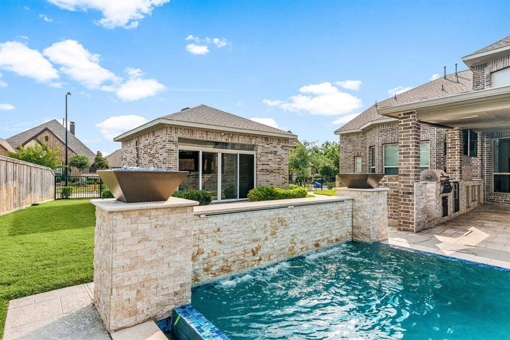 This photo showcases a modern backyard with a swimming pool featuring a waterfall edge. The area is accented by stone and brickwork, and includes a built-in outdoor kitchen. The well-maintained lawn and privacy fence complete the inviting outdoor space.