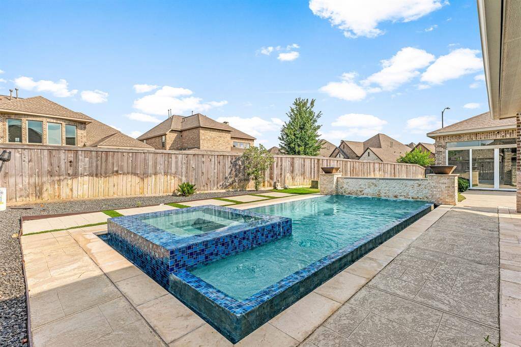 This photo showcases a modern backyard with a sleek pool, featuring a raised hot tub area with blue mosaic tiles. The space is bordered by a wooden privacy fence and includes a paved patio area, creating a stylish and functional outdoor retreat.