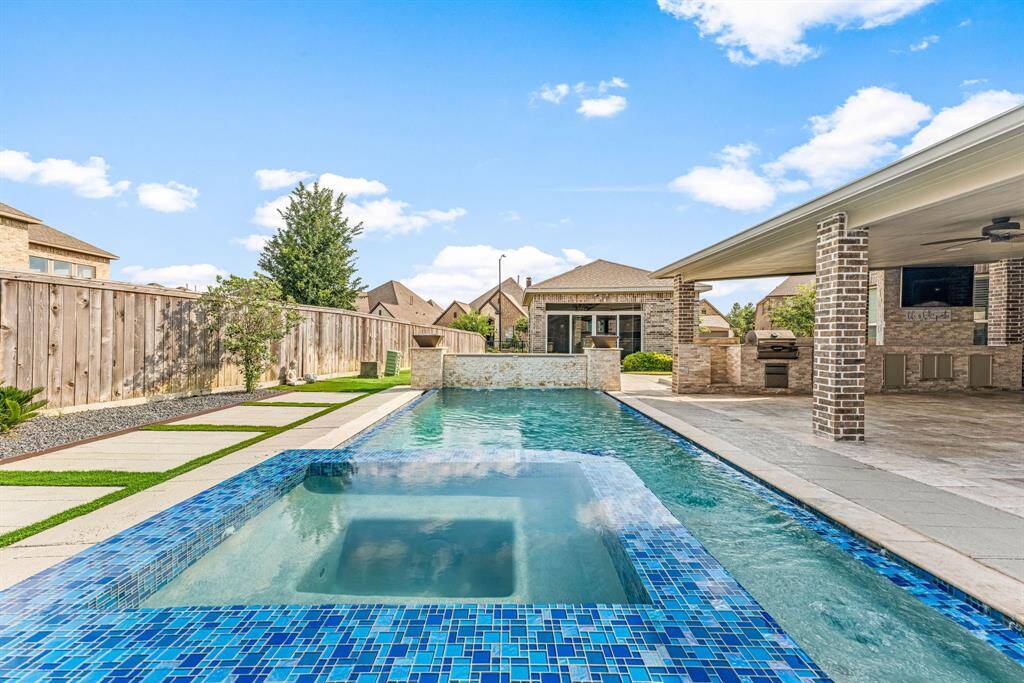 This photo showcases a luxurious backyard with a modern swimming pool featuring a hot tub. The area is surrounded by clean landscaping and a privacy fence. The covered patio includes an outdoor kitchen, perfect for entertaining.