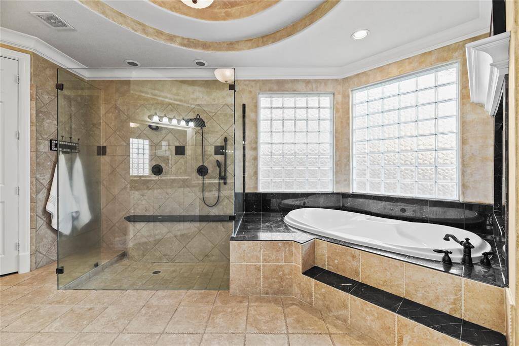 Glass block windows bathes the space in soft, diffused light, while the expansive walk-in shower features dual rain shower heads for an indulgent experience. A grand Roman-inspired step-up soaking tub offers a serene retreat, perfectly blending classical elegance with modern luxury.