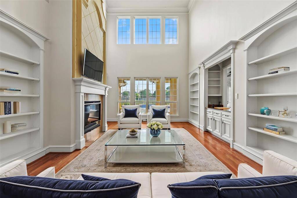 The formal sitting area captivates with soaring two-story ceilings and an abundance of natural light that pours through expansive windows. A striking central fireplace serves as the focal point, flanked by custom-built white cabinetry that adds a touch of timeless elegance and sophistication.