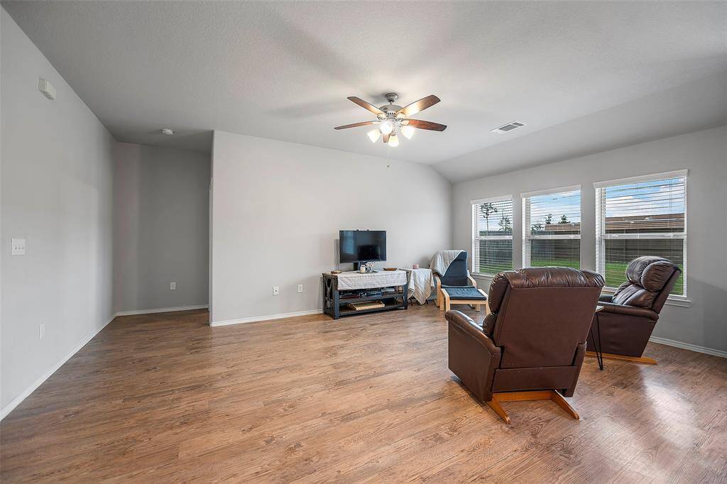 This is a well-lit, spacious living room with laminate wood flooring, a ceiling fan, and large windows offering plenty of natural light. Plenty of space to add an oversized sectional or a sofa and chairs.