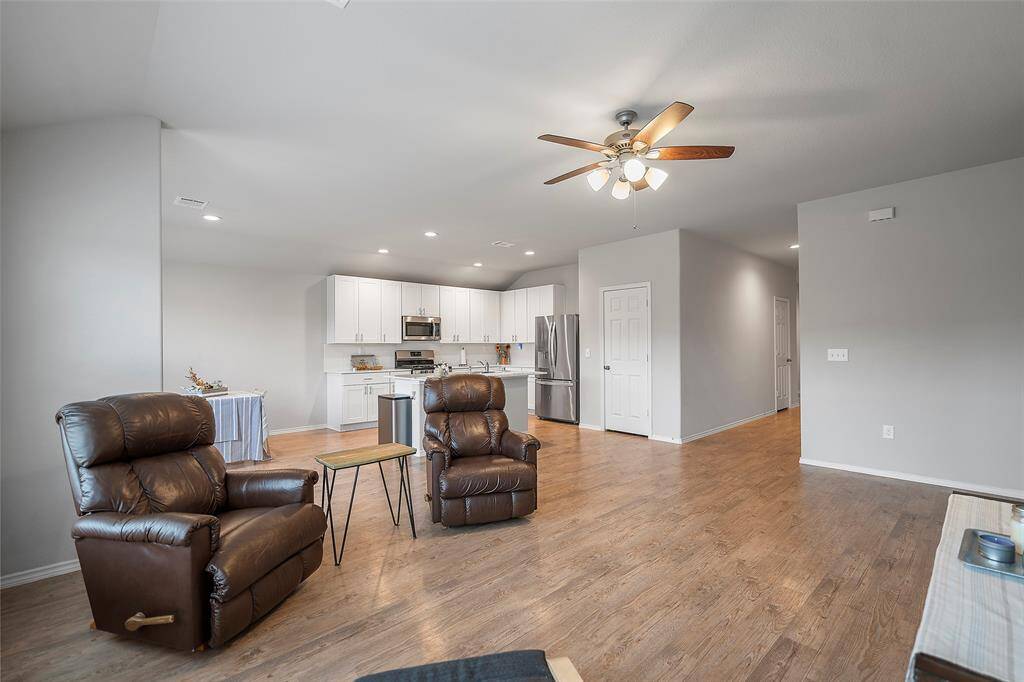 The photo shows a modern open-concept living space with a connected kitchen, featuring white cabinetry and stainless steel appliances. The area is well-lit with both natural and ceiling fan lights, and has laminate wood flooring throughout.