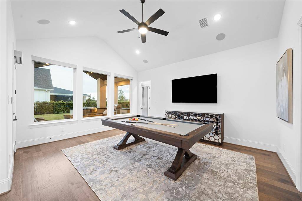 The game room balances comfort and style with a vaulted ceiling, warm hardwoods, and large windows that capture serene backyard views and covered patio access.