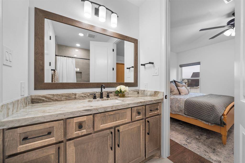 The secondary ensuite bath showcases thick stone counters, a wide framed mirror, and custom cabinetry built for everyday utility.