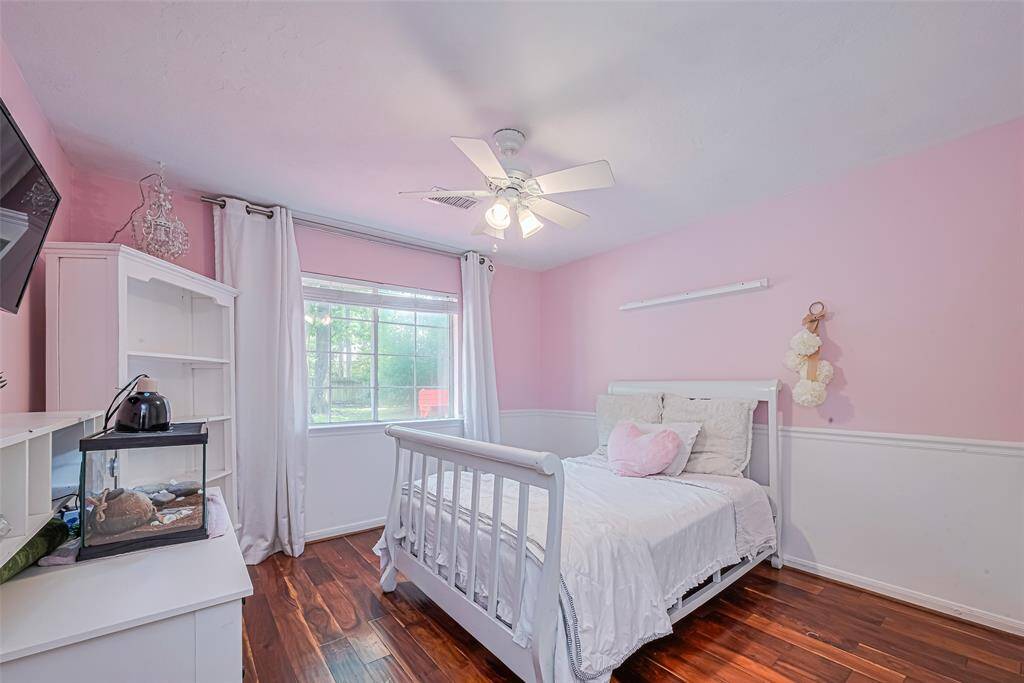 This bedroom features soft pink walls with white wainscoting, a large window for natural light, a fan to cool and warm flooring. A view of the pool is from the window and creating a charming and inviting space.