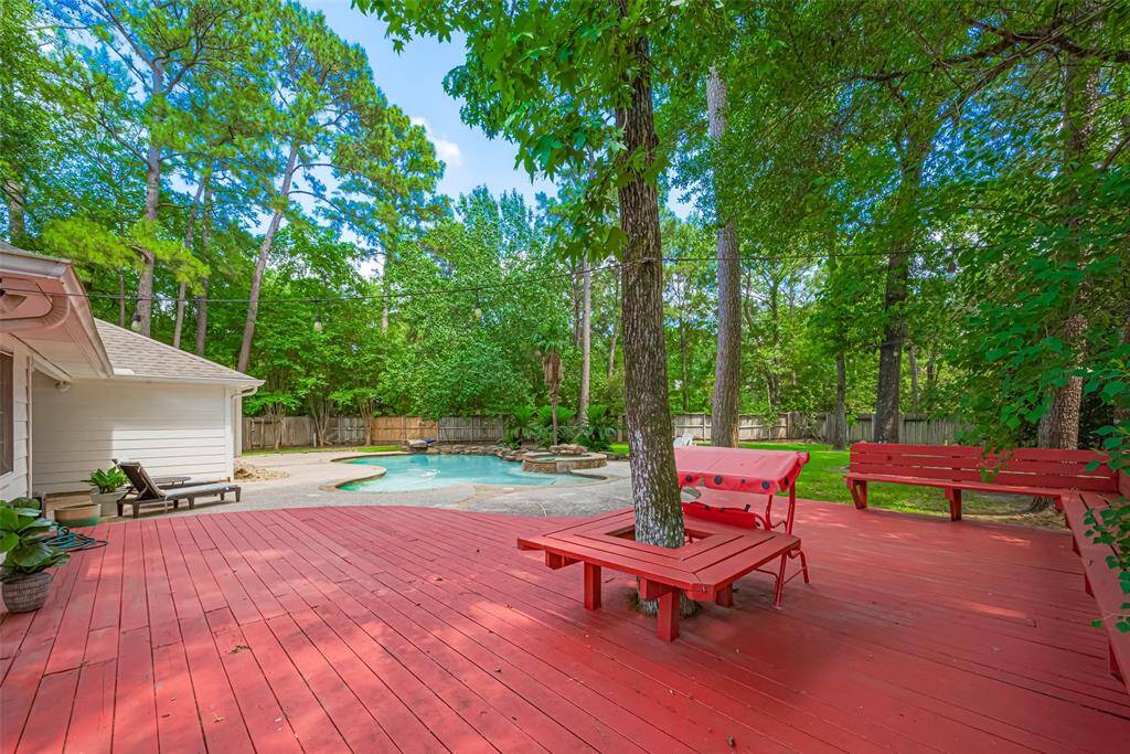This backyard features a spacious red deck with built-in seating, shaded by mature trees. A pool with a natural-style design is surrounded by a lush, wooded fence line, offering privacy and a tranquil setting. Ideal for outdoor relaxation and entertaining.