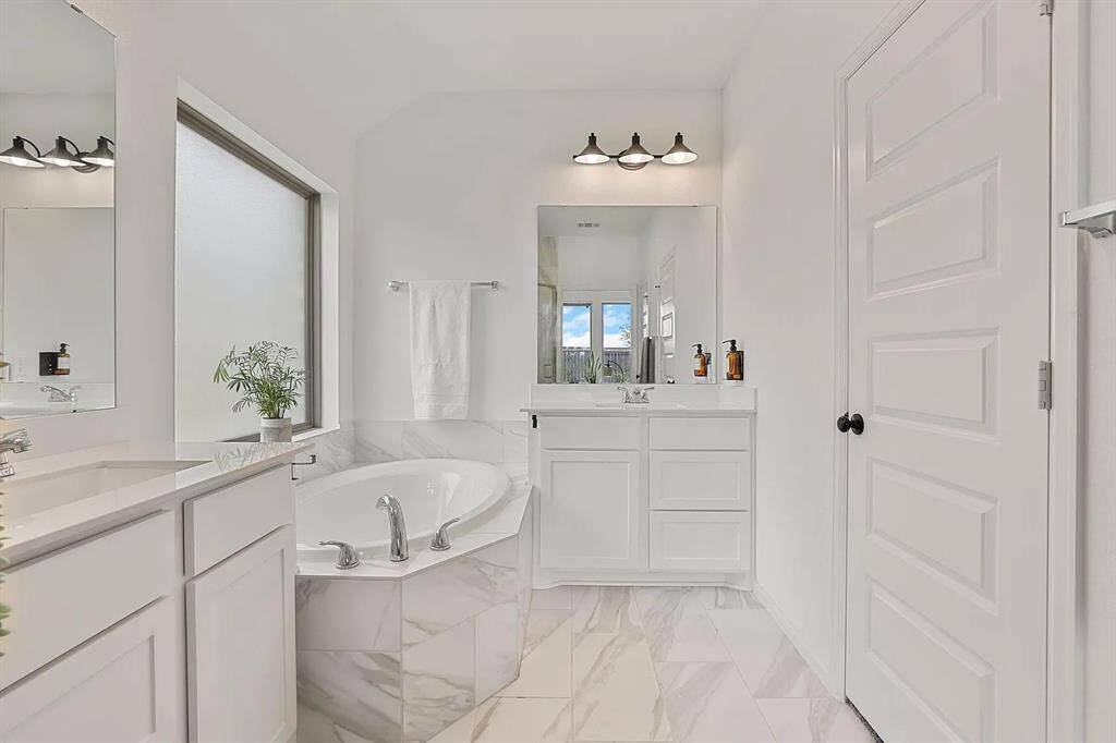 This bathroom features a modern design with a large soaking tub, elegant white cabinetry, and a spacious vanity with ample lighting. The frosted window offers natural light while maintaining privacy, and the sleek marble-like flooring adds a touch of luxury.