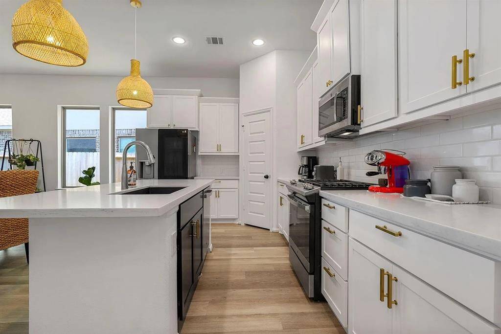 This modern kitchen features sleek white cabinetry with gold hardware, stainless steel appliances, and a spacious island with a black sink. The room is brightened by stylish pendant lights and natural light from large windows, complemented by light wood flooring.